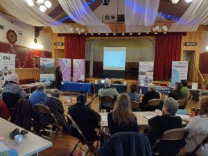 A group of people attending the 'Community action for a greener future' event. Various pop-up banners are on either side of the stage, and slides are being projected onto a pull-down white projector screen.
